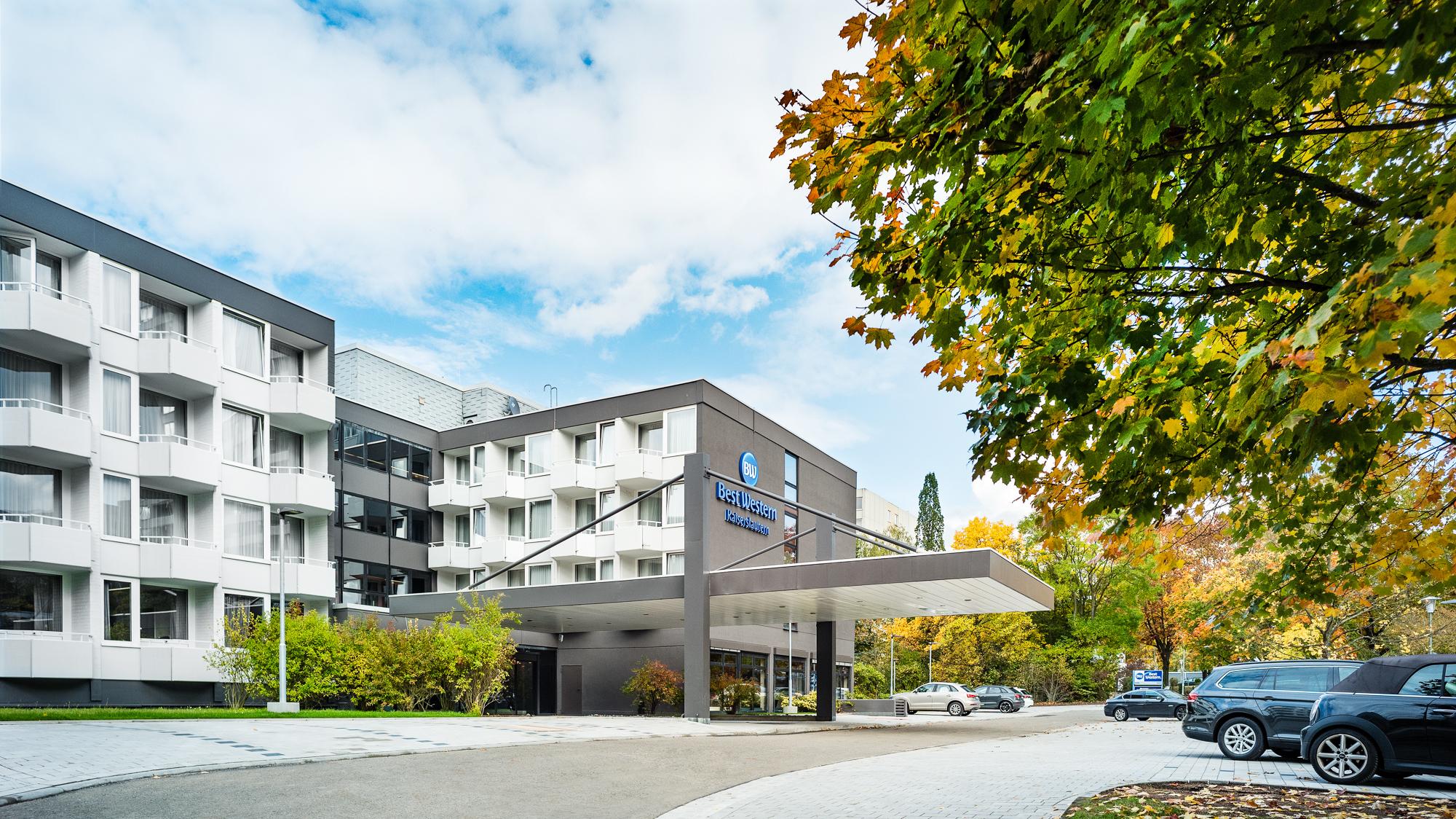 Moderne Best Western Hotel-Fassade mit Balkonen, großem Logo, überdachtem Eingang und geparkten Autos bei teils bewölktem Himmel.