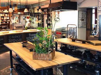 Moderne Küche mit Holztischen, schwarzen Regalen, hängenden Utensilien und Kreidetafeln im Hintergrund.