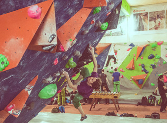 Boulderhalle mit Kletternden, Fokus auf Person mit lila Shirt und grüner Hose an überhängender Wand mit bunten Griffen.