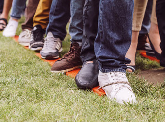 Reihe von Personen auf Grasfläche, Füße entlang einer orangen Linie, verschiedene Schuhe wie Sneaker, Sandalen und Freizeitschuhe.