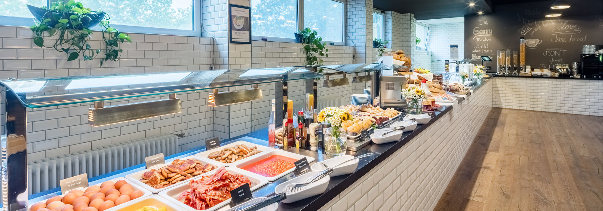 Buffet mit Frühstücksgerichten wie Eiern, Würstchen, Bohnen, Speck unter Wärmelampen, dahinter Müsli und Sprüche an der Wand.