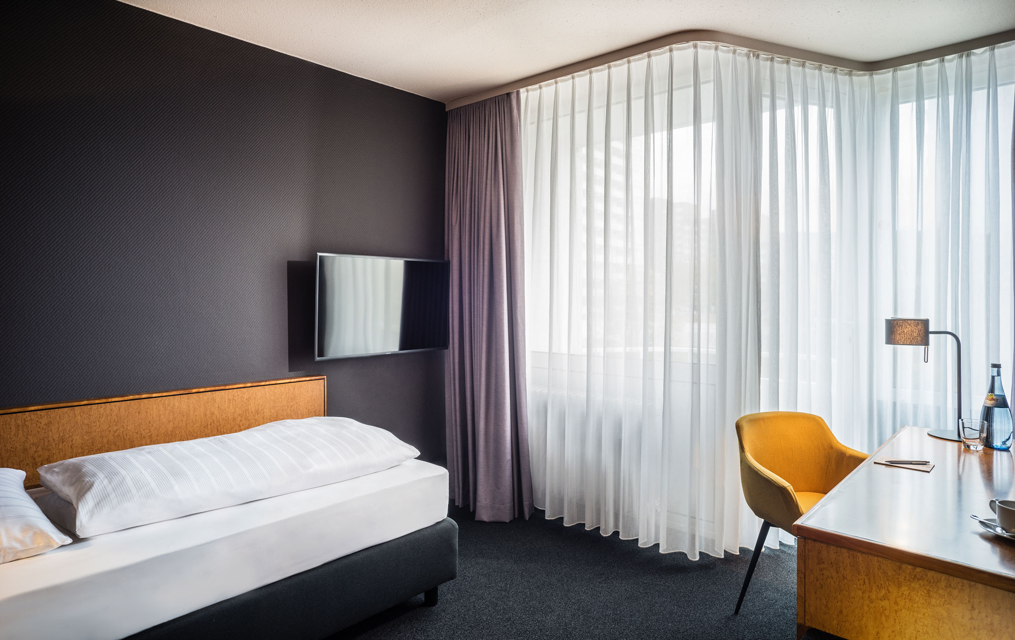 Modernes Hotelzimmer mit Einzelbett, Wand-TV, Schreibtisch mit orangefarbenem Stuhl, großer Fensterfront und Tageslicht.