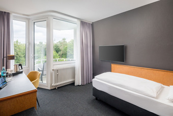 Modernes Schlafzimmer mit Einzelbett, Schreibtisch, Stuhl, großem Fenster, grauen Wänden, Teppichboden und Wand-TV.