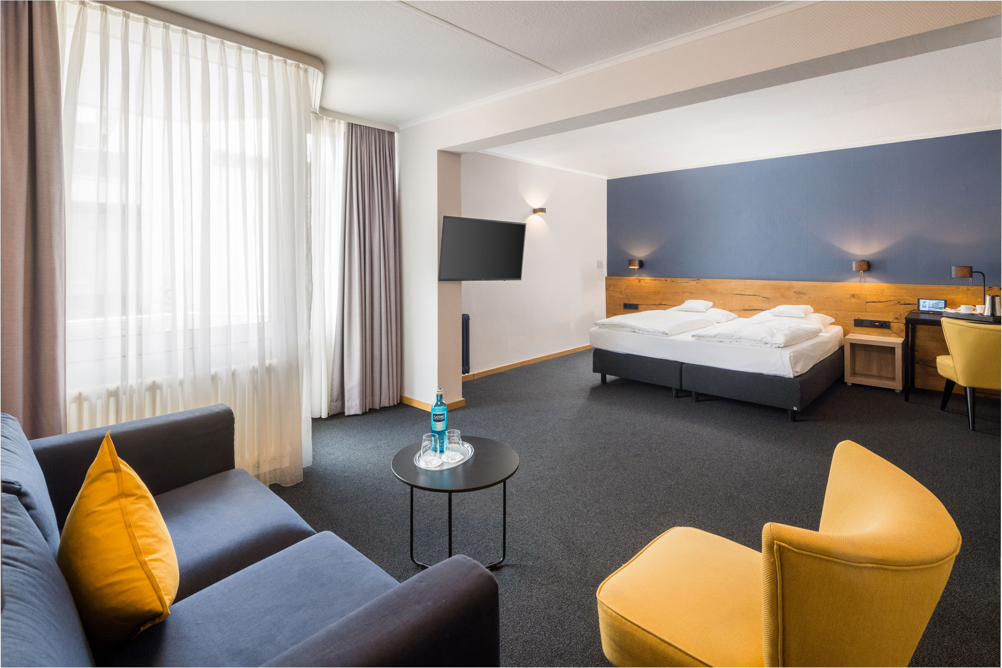 Hotelzimmer mit Doppelbett, Sofa, gelben Sesseln, Tisch mit Wasserflasche, Schreibtisch und Fernseher
