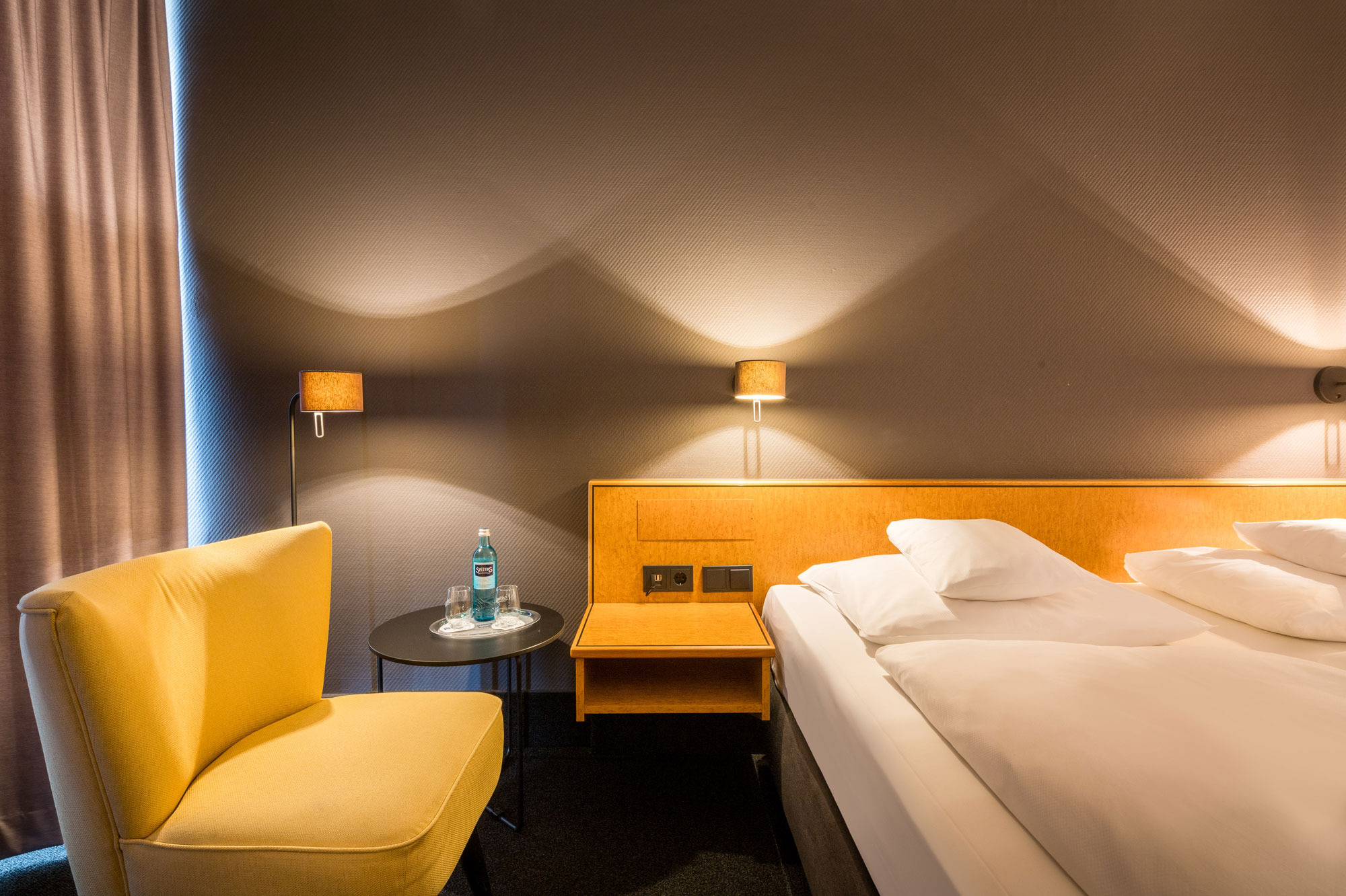 Modernes Hotelzimmer mit Bett, gelbem Stuhl, Wandlampen, kleinem Tisch mit Wasserflasche und warmem Licht.