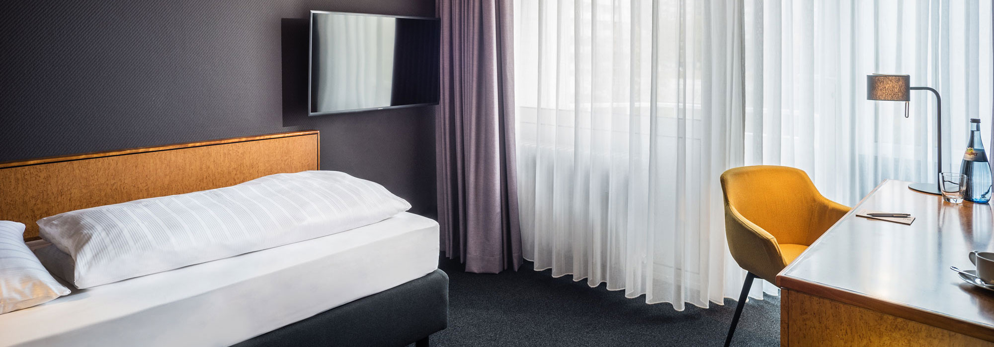 Modernes Hotelzimmer mit Einzelbett, Wand-TV, Schreibtisch mit orangefarbenem Stuhl, großer Fensterfront und Tageslicht.