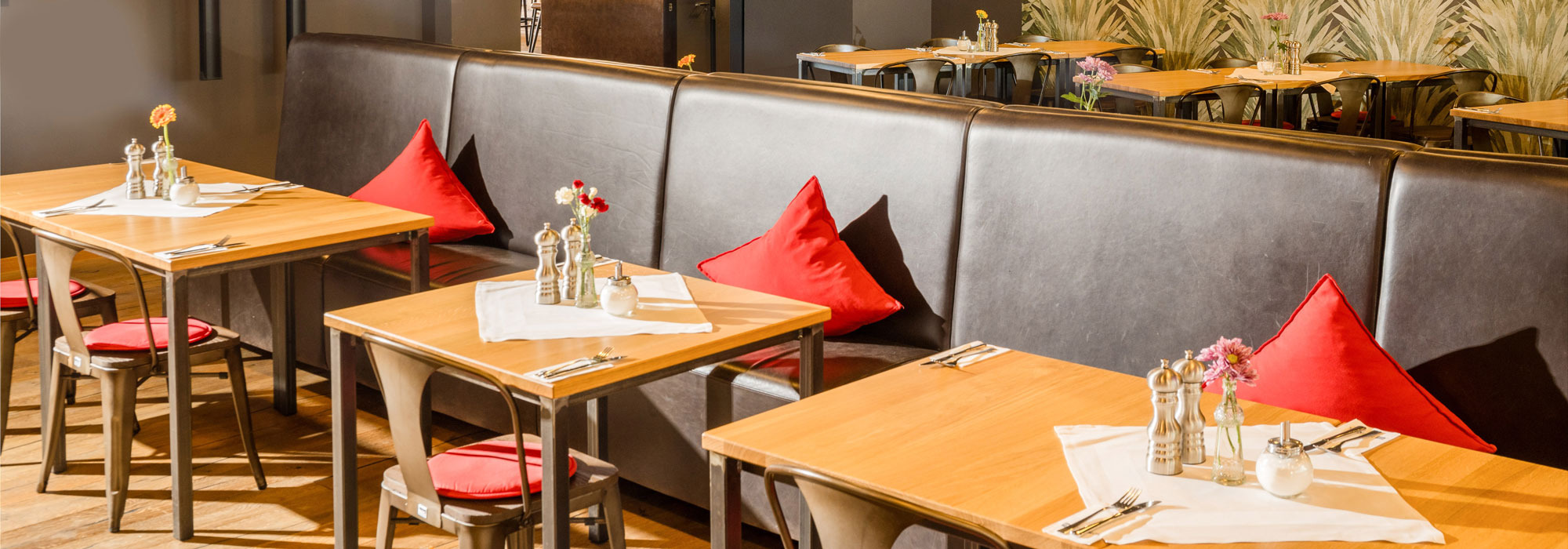 Gemütliches Restaurant mit Holztischen, weißen Tischdecken, Blumenvasen, roten Kissen und dekorativer Wandgestaltung.
