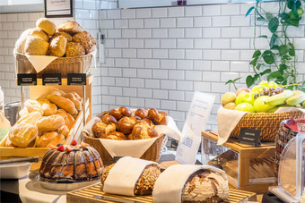 Auswahl an Brötchen, Brezeln, Kuchen und Obst auf Theke mit Beschriftungen vor weiß gefliester Wand mit Pflanzen.