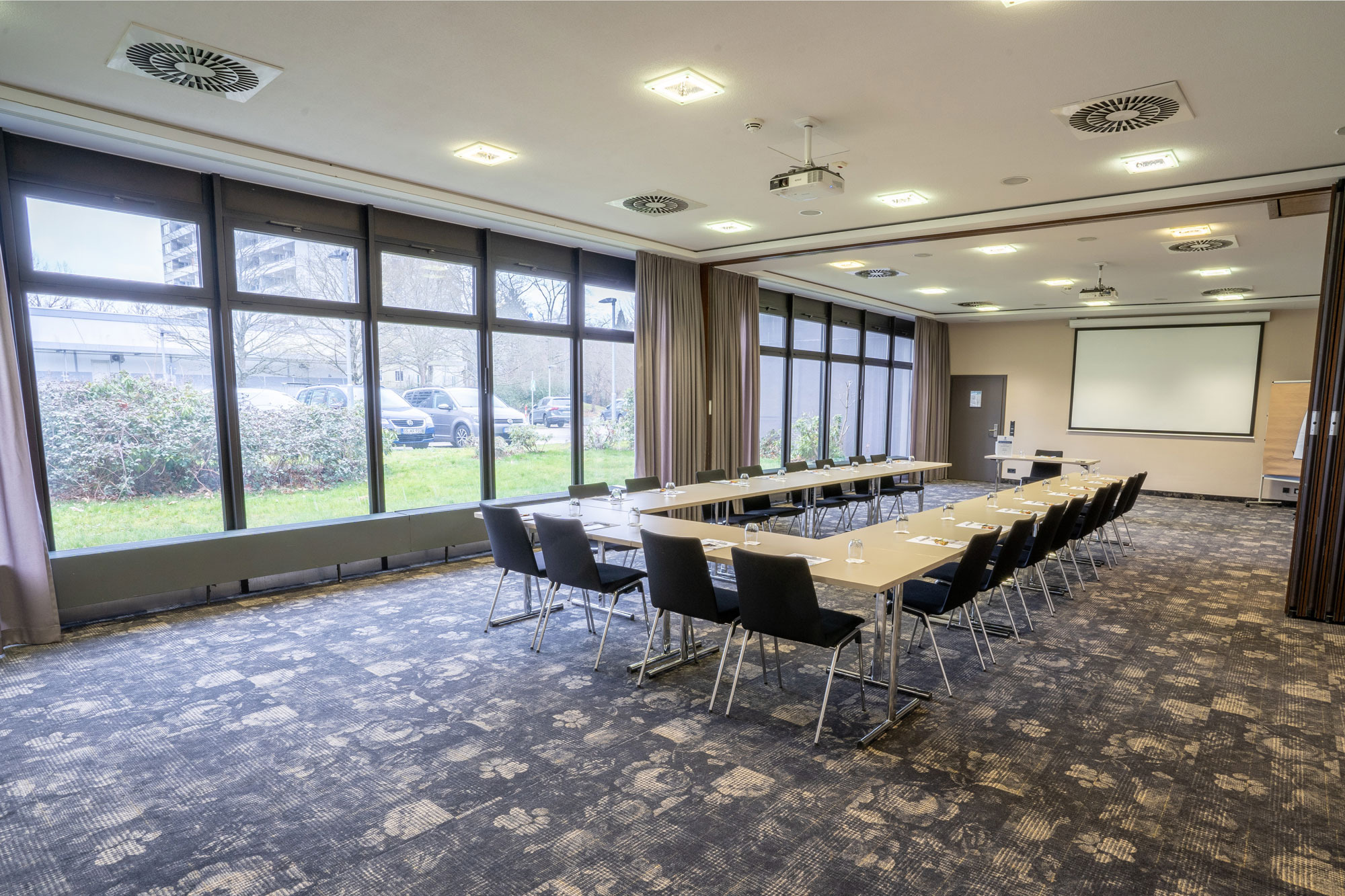 Konferenzraum mit U-förmigem Tisch, Stühlen, Wasser, Schreibmaterial und Projektionsfläche, Tageslicht von Fensterseite.