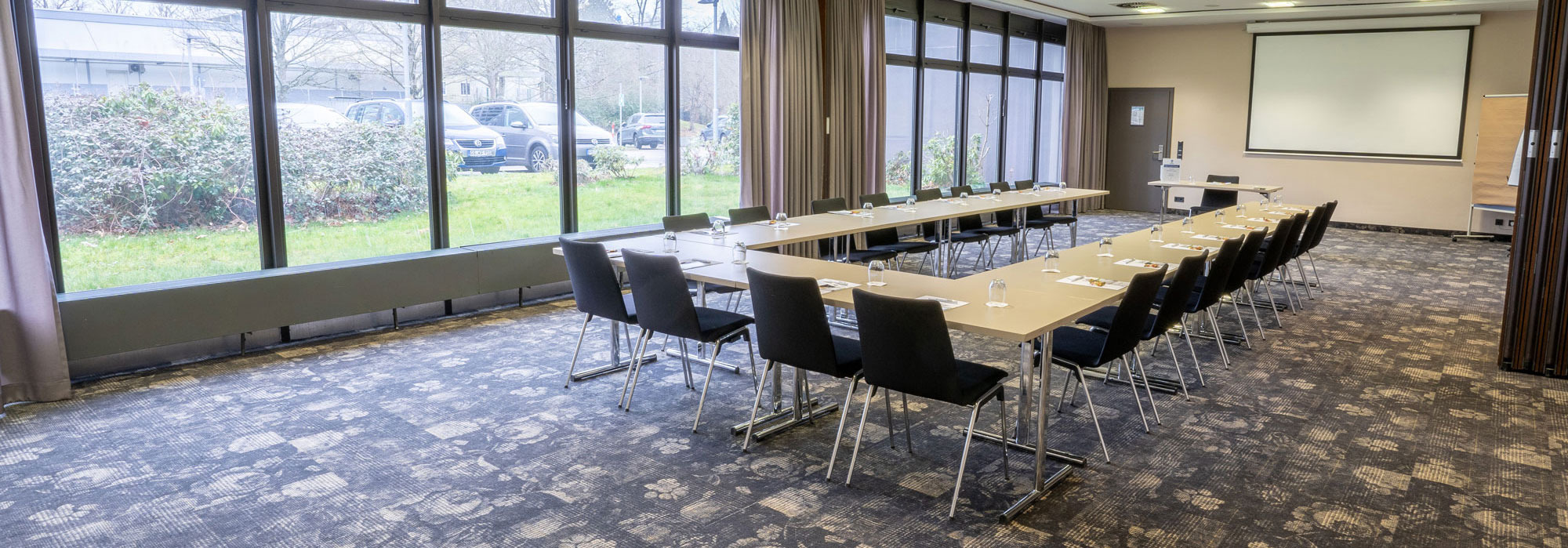 Konferenzraum mit U-förmigem Tisch, Stühlen, Wasser, Schreibmaterial und Projektionsfläche, Tageslicht von Fensterseite.