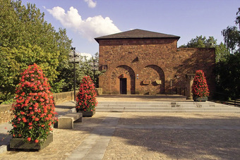 Casimirschloss aus rotem Backstein mit Rundbögen, Innenhof mit Pflanzkübeln, Bänken und Bäumen bei teils bewölktem Himmel.