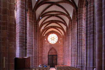 Innenraum einer Kirche mit hohen Gewölbedecken, roten Steinsäulen, Rundfenster mit Buntglas und Stuhlreihen im Kirchenschiff.