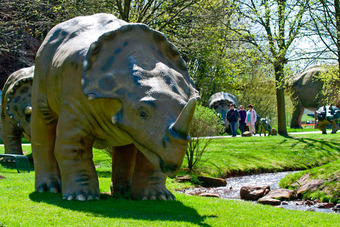 Park mit lebensgroßen Dinosaurier-Skulpturen, im Vordergrund ein Triceratops nahe einem Bach, umgeben von Bäumen und Spaziergängern.