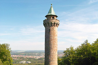 Humbergturm mit grünem Spitzdach, umgeben von Bäumen, mit Blick auf eine Stadt im Hintergrund unter leicht bewölktem Himmel.