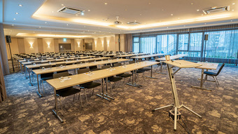 Großer Konferenzraum mit Reihen von Tischen und Stühlen, Teppichboden, Tageslicht und heller Deckenbeleuchtung.